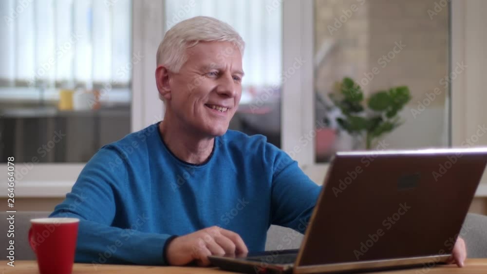 Closeup shoot of aged caucasian man typing on the laptop smiling happily indoors in a cozy apartment Closeup shoot of aged caucasian man typing on the laptop smiling happily indoors in a cozy apartment