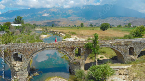 AERIAL: Woman in bright red dress strolling along the decaying arched bridge.