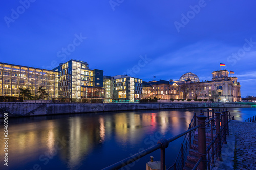 Illuminated modern and old parliament buildings including the historic Reichstag and their reflections on the Spree River in Berlin, Germany, at dusk.