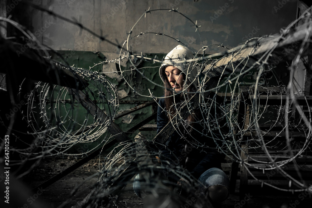 Young girl is sitting behind barbwire in old, ruined factory.