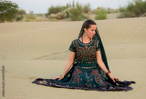 A young woman in a national Indian costume sits thoughtfully on the sand among the dunes in the Thar desert. Rajasthan India.