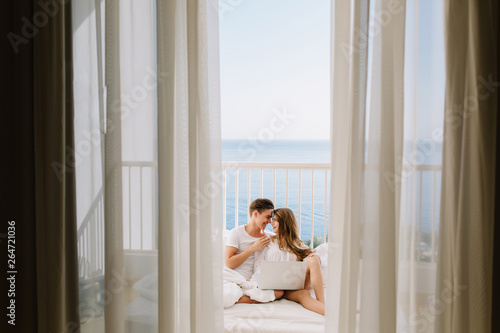 Portrait of loving couple watching movie together on balcony in morning with curtains on foreground. Young man resting with his amazing tanned girlfriend in bed holding laptop with sea background