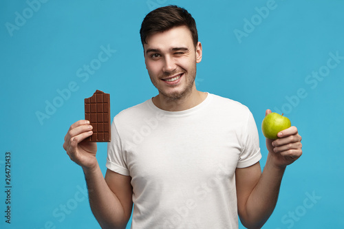 Joyful young Caucasian brunette guy in white t-shirt made wrong choicem deciding between milky chocolate and fresh green apple. People, health, food, enjoyment, dieting and calories concept