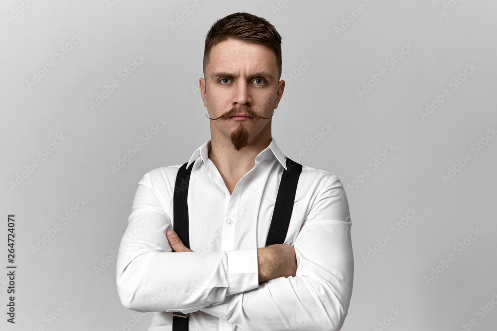 Stylish confident bearded boss in suspenders and white shirt posing isolated at blank copyspace studio wall, crossing arms on his chest, angry with inefficient business strategy, having strict look Stylish confident bearded boss in suspenders and white shirt posing isolated at blank copyspace studio wall, crossing arms on his chest, angry with inefficient business strategy, having strict look