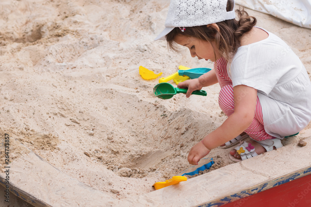 Little caucasian girl in a cap, t-shirt and striped leggings playing in the sandbox on the playground, close-up, day Little caucasian girl in a cap, t-shirt and striped leggings playing in the sandbox on the playground, close-up, day