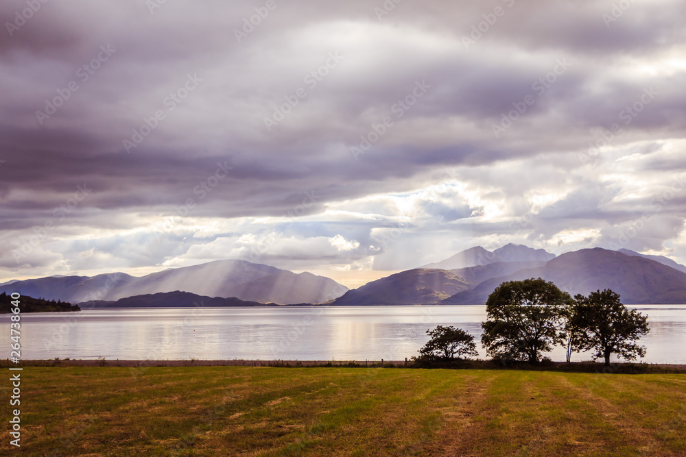 Mystic landscape lake scenery in Scotland: Cloudy sky, meadow, trees and lake with sunbeams, mountain range in the background. Loch Linnhe.