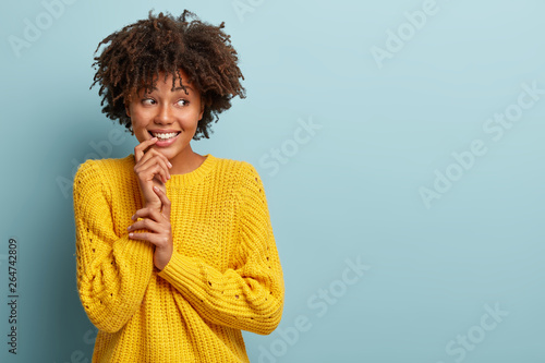 Optimistic lovely woman with curly hair, keeps hand near toothy smile, looks aside, has dreamy expression, feels shy, wears yellow casual jumper, stands against blue background. Copy space right © Wayhome Studio