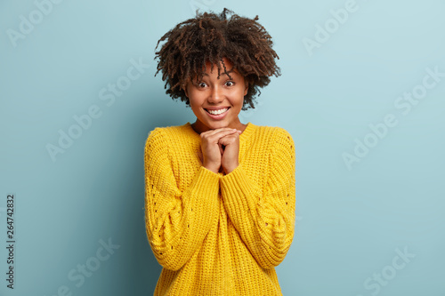 Touched pleased Afro American woman holds hands together under chin  feels admiration  expresses positive emotions  has toothy smile  wears yellow jumper  has fun and laughs indoor. Emotions concept