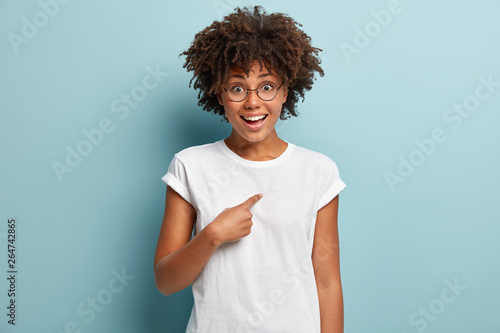 Select me please. Joyous dark skinned female points finger at herself, can not believe her luck, wears casual t shirt, isolated over blue studio wall, says I am what you need. Me is best option