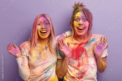 Holi festival, fun and people concept. Two overjoyed women play with colors, show palms, smile gladfully, smeared with colorful powder, isolated over purple background. Multicolored explosion