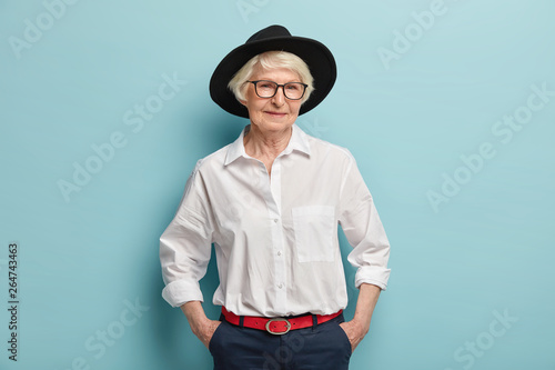 Fashionable mature woman wears stylish black hat, white shirt and trousers with red belt, keeps hands in pockets, has glad expression, isolated over blue background. People, age and fashion concept