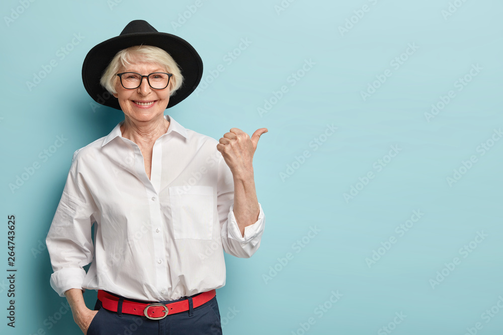 Waist up shot of friendly looking senior lady in stylish headgear, white elegant shirt and formal trousers, holds hand in pocket, points thumb away, has happy smile, advertises something nice - obrazy, fototapety, plakaty 