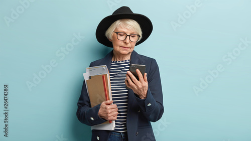 Bild auf Leinwand Concentrated elderly headmistress focused in cell phone, holds some business pap