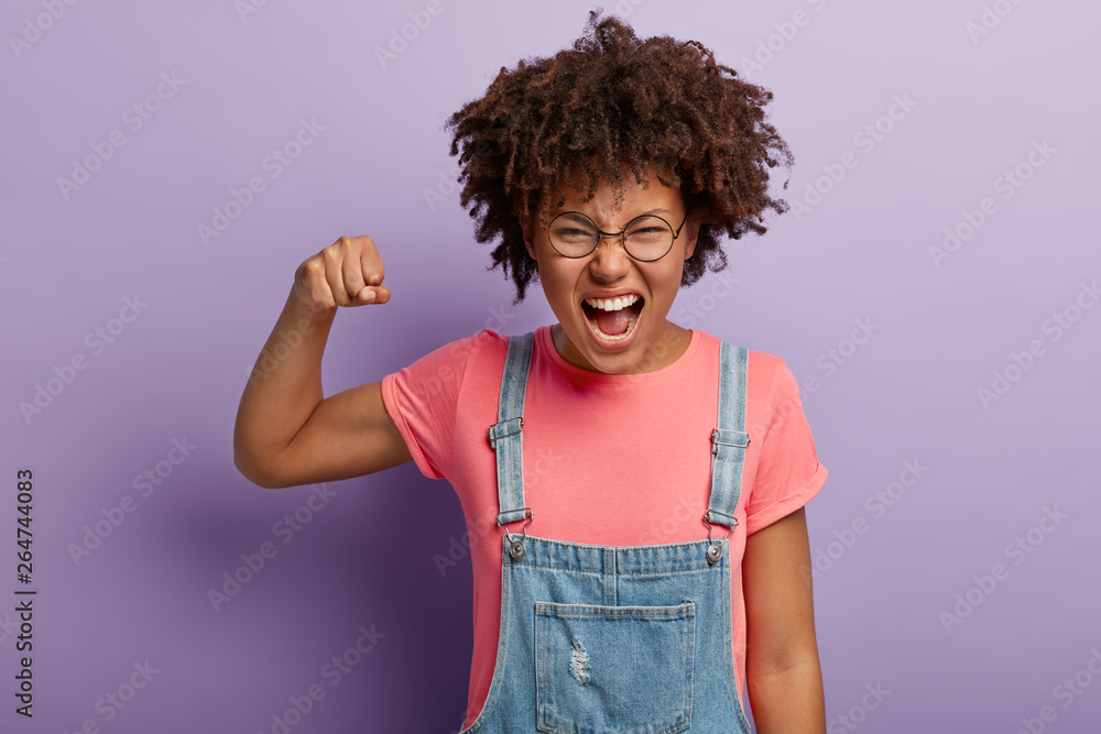 Emotional African American girl raises fist, exclaims from positive emotions, smirks face, dressed in pink t shirt and denim sarafan, stands against purple background. We did together, congratulation! Emotional African American girl raises fist, exclaims from positive emotions, smirks face, dressed in pink t shirt and denim sarafan, stands against purple background. We did together, congratulation!