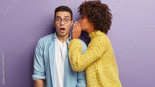 Photo of Afro American woman whispers secret to boyfriend, tells shocking news. Astonished young man keeps jaw dropped from surprisement, hears terrible rumors, isolated over purple background