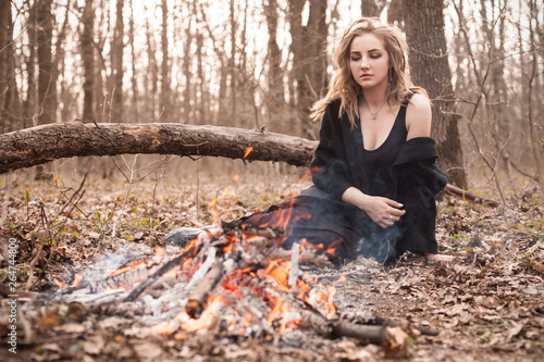 Young woman with black cape sitting in forest near bonfire.