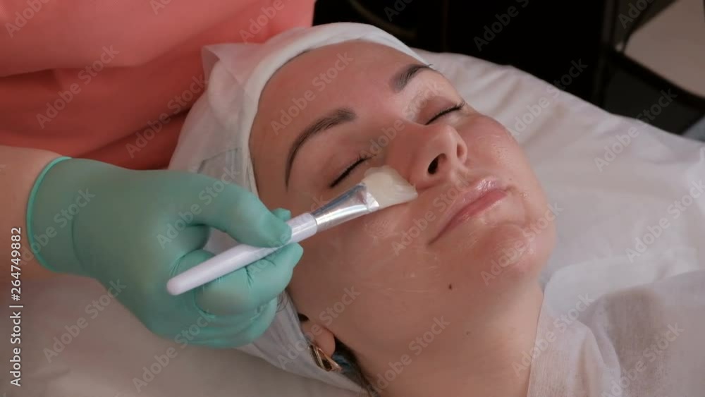 Beautiful European girl with closed eyes on the procedure in the beauty salon. Beautician hands in green gloves holding a white brush. Applying a transparent nutritional gel on a woman's face. Beautiful European girl with closed eyes on the procedure in the beauty salon. Beautician hands in green gloves holding a white brush. Applying a transparent nutritional gel on a woman's face.