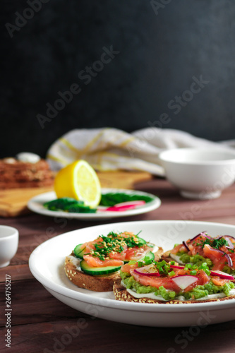 open sandwiches with salmon cucumber mashed avocado red onion dill on white plate and ingredients on the table close up soft focus vertical orientation