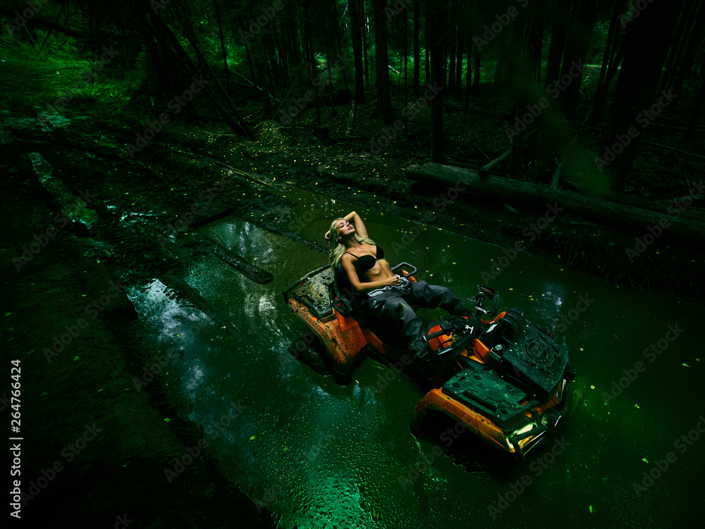 Beautiful blonde girl on a orang ATV rides on the river, standing on the mountain. Around the forest, sand and blue sky.She rides on a green swamp, in the mud and all wet. She's wearing a blue bikini. Beautiful blonde girl on a orang ATV rides on the river, standing on the mountain. Around the forest, sand and blue sky.She rides on a green swamp, in the mud and all wet. She's wearing a blue bikini.