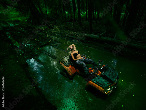 Beautiful blonde girl on a orang ATV rides on the river, standing on the mountain. Around the forest, sand and blue sky.She rides on a green swamp, in the mud and all wet. She's wearing a blue bikini.