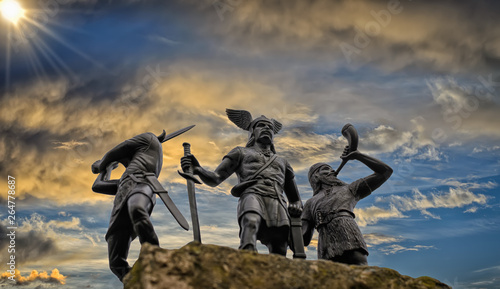 Old Norse God Odin on a rock, flanked by two Vikings (toy soldiers), one of them raised a sword and the other is trumpeting with horn, blue sky, bright sun, dramatic clouds