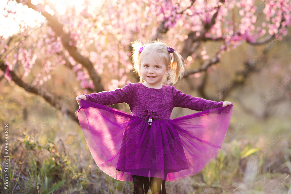 Pretty baby girl 1-2 year old wearing princess dress poaing over blooming tree outdoors. Looking at camera. Spring season.