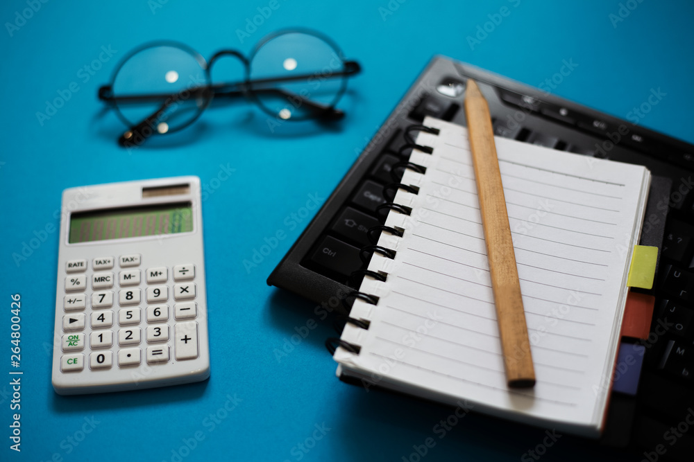 Top view of blue office desk, with pencil and empty notebook on keyboard, glasses and white calculator. Top view of blue office desk, with pencil and empty notebook on keyboard, glasses and white calculator.