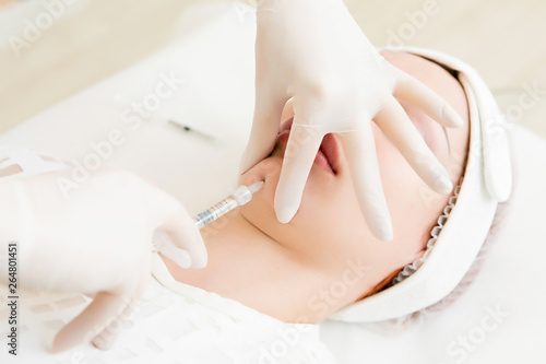 a girl in a white sterile hat is lying on a couch by a beautician and she is given injections for a skin tightening on her chin.