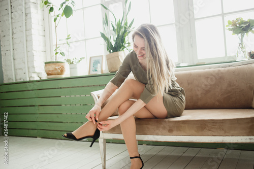 Girl in a short khaki dress buttons her sandals before photoshoot and party. Beautiful slender legs.