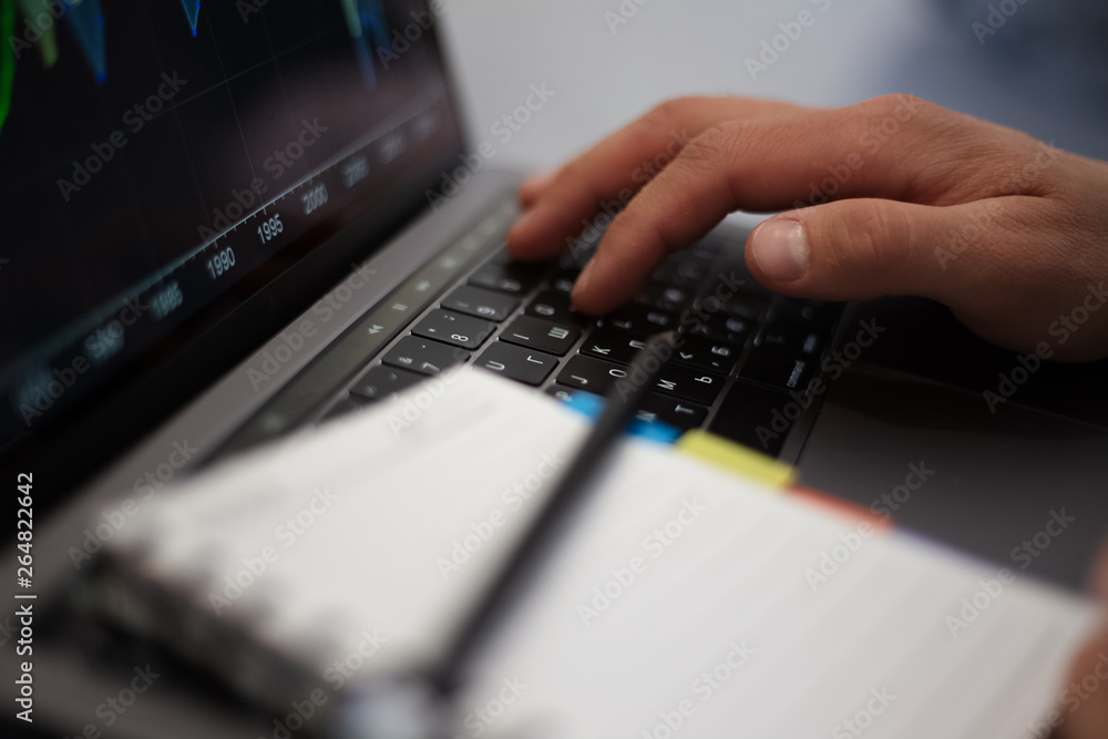 Close-up of man hands, typing on keyboard of laptop and holding mug of coffee, near notebook with black pencil.