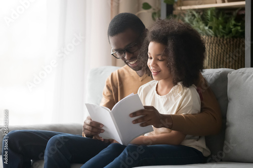 Black father reading book to daughter sitting on couch