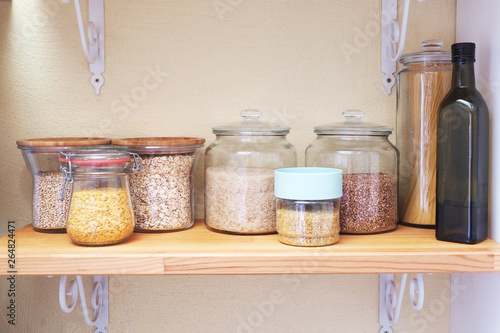 Glass jars with products and olive oil are on a wooden shelf.
