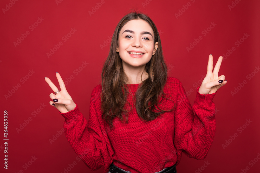 Portrait of gorgeous woman looking at camera with smile and showing peace sign with fingers isolated over red background Portrait of gorgeous woman looking at camera with smile and showing peace sign with fingers isolated over red background