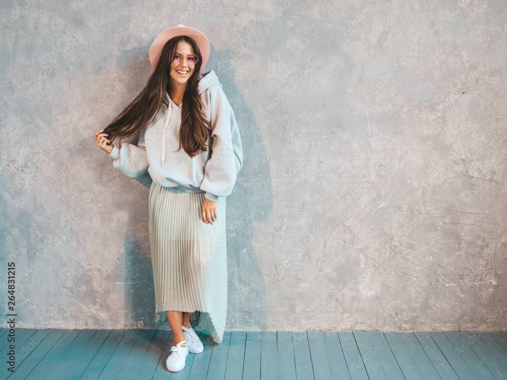 Young beautiful smiling woman looking at camera. Trendy girl in casual summer hoodie and skirt clothes. Funny and positive female posing near gray wall in studio. In hat Young beautiful smiling woman looking at camera. Trendy girl in casual summer hoodie and skirt clothes. Funny and positive female posing near gray wall in studio. In hat