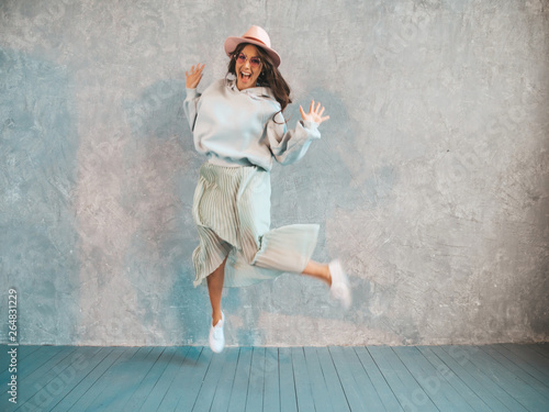Beautiful cute smiling model jumping. Girl in summer hipster hoodie and skirt. Model having fun and going crazy near gray wall in studio