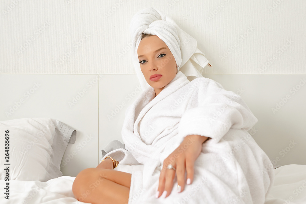 A young girl with white hair in her bedroom is posing on a bed in a white coat and a white towel on her head. A young girl with white hair in her bedroom is posing on a bed in a white coat and a white towel on her head.