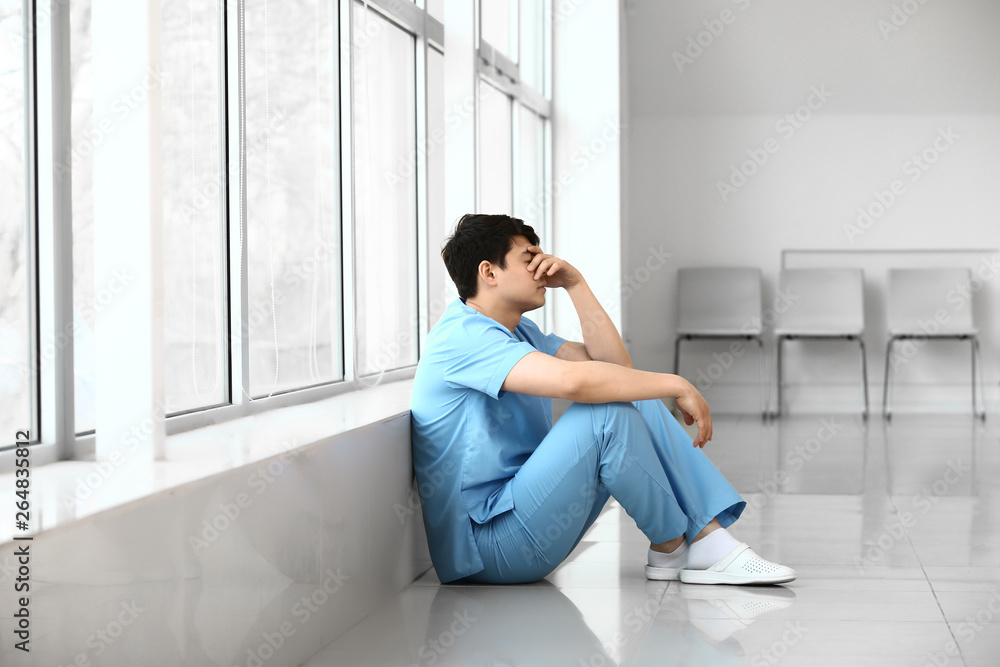 Depressed male medical assistant sitting near window in clinic Depressed male medical assistant sitting near window in clinic