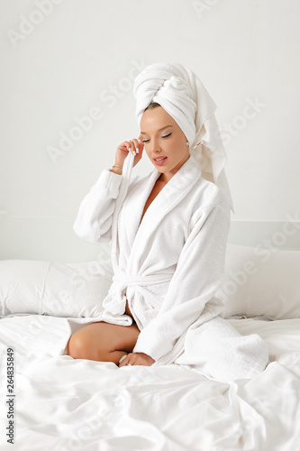 A young girl with white hair in her bedroom is posing on a bed in a white coat and a white towel on her head.