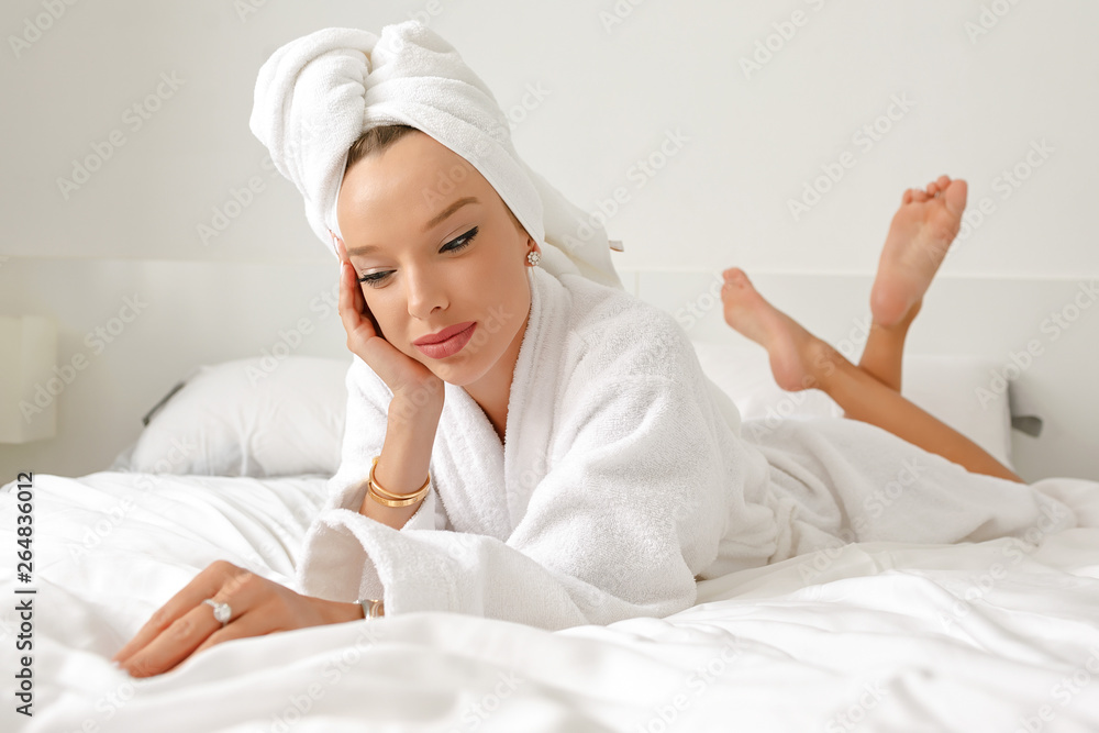 A young girl with white hair in her bedroom is posing on a bed in a white coat and a white towel on her head. A young girl with white hair in her bedroom is posing on a bed in a white coat and a white towel on her head.