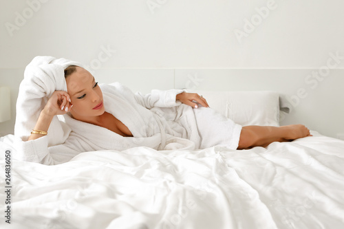 A young girl with white hair in her bedroom is posing on a bed in a white coat and a white towel on her head.