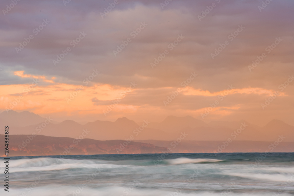 Dreamy blurred waves under an orange and purple sunset sky in Plettenberg Bay, with mountains in the distance. Garden Route, Western Cape, South Africa