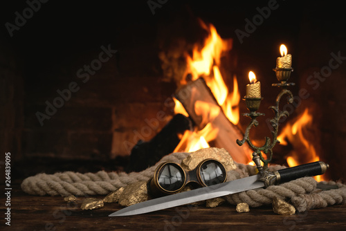 Pirate concept. Treasure hunt. Adventurer table with treasure gold, sword, binoculars and rope on a burning fire background.