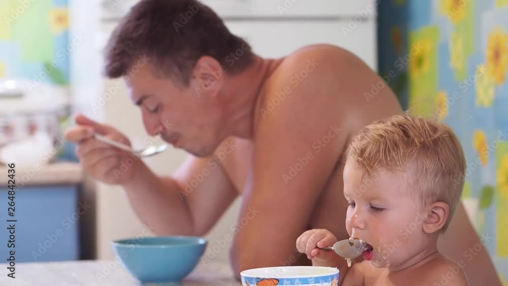 Dad and baby boy sit in the kitchen and eat food together The best dad in the world is sitting in the kitchen at the table with a little boy of European appearance and eat food in a place. Dad and baby boy sit in the kitchen and eat food together The best dad in the world is sitting in the kitchen at the table with a little boy of European appearance and eat food in a place.