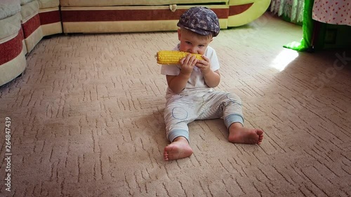 Charming attractive cute baby in hat sitting on the floor emotionally enjoying juicy boiled corn 
