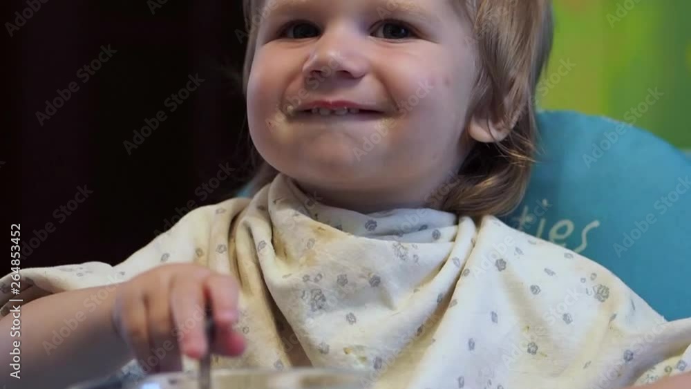 A little boy sitting at a table and eating Blonde child of European appearance while in the kitchen holding a spoon sitting at the table and emotionally eating
