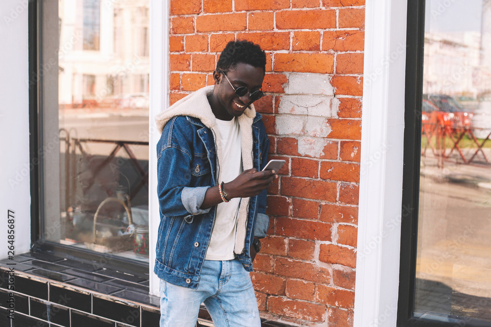 Stylish cool smiling african man with smartphone on city street over brick wall background