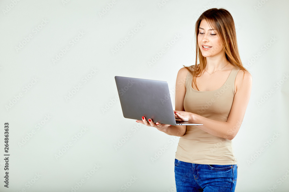 a girl in a beige t-shirt and blue jeans is standing against a gray wall holding a laptop and is looking at his screen smiling