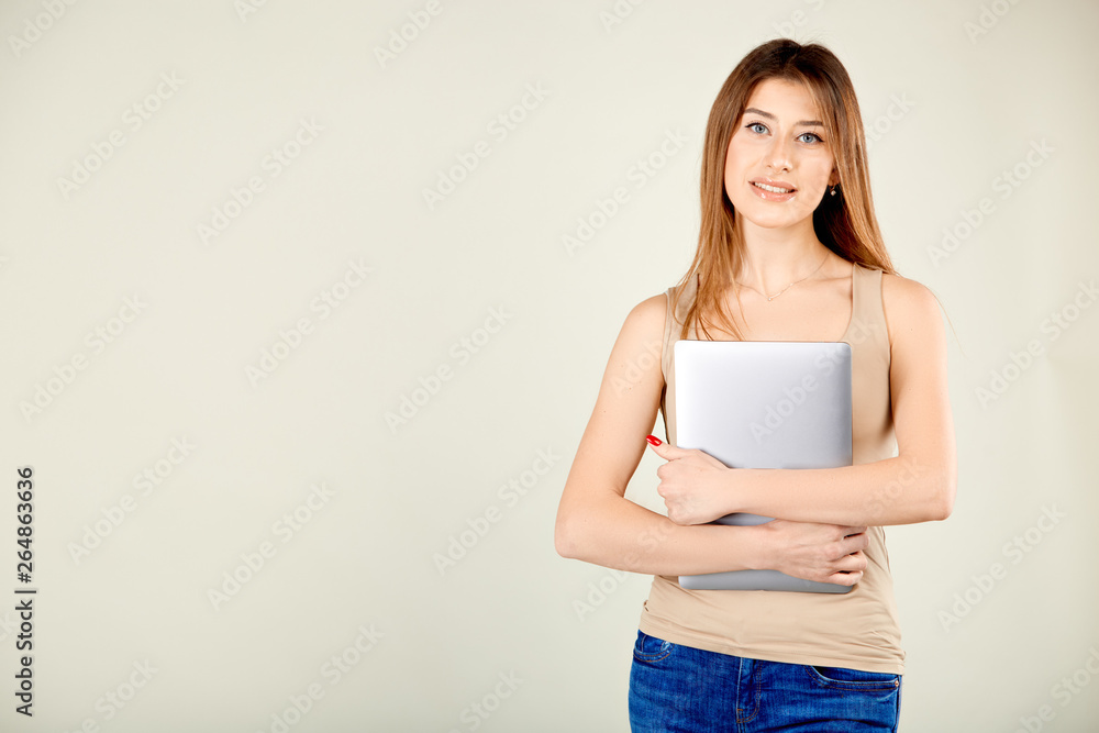 a girl in a beige t-shirt and blue jeans is standing against a gray wall holding a closed laptop in her hands a girl in a beige t-shirt and blue jeans is standing against a gray wall holding a closed laptop in her hands