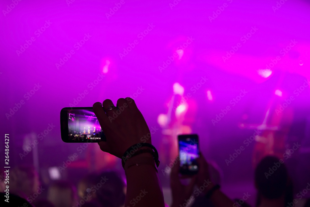 Man hand with a smartphone records a live concert of the group consisting of four cellists and a drummer. The scene is lit with purple spotlights Man hand with a smartphone records a live concert of the group consisting of four cellists and a drummer. The scene is lit with purple spotlights