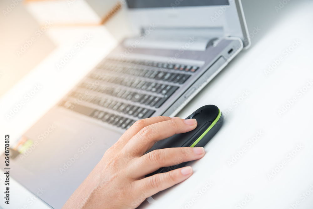 Fototapeta premium Closeup woman's hands clicking mouse and working on a laptop computer on a white desk with sun flare lighting effect .